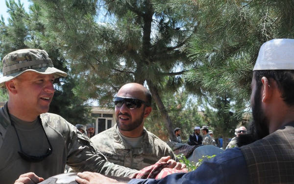 Two U.S. Army soldiers in MultiCam/OCP camouflage uniforms interact with local civilians, appearing to examine or treat a dark-colored horse or large animal. The setting appears to be an outdoor area in what looks like Afghanistan, with pine trees and a tan/adobe-style building visible in the background. The interaction suggests a veterinary civil affairs or humanitarian assistance activity.