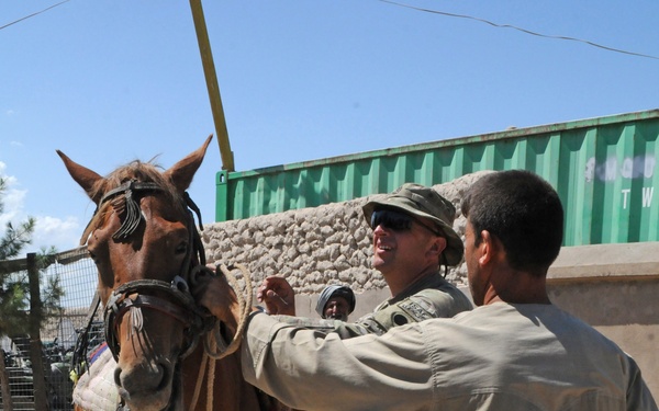 A uniformed military personnel wearing a patrol cap and sunglasses interacts with a local civilian while both attend to a chestnut horse with traditional decorative bridle. The scene takes place in an arid outdoor setting with a stone wall and green shipping container visible in the background. The interaction appears cooperative and friendly in nature.