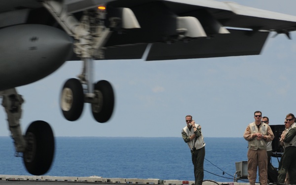 A military jet aircraft is captured in the foreground with its landing gear deployed, appearing to be on approach or just landing on an aircraft carrier flight deck. Several flight deck personnel in tan/khaki flight suits and safety vests are standing and observing near a deck-edge control station. The ocean horizon is visible in the background, confirming the carrier is at sea.