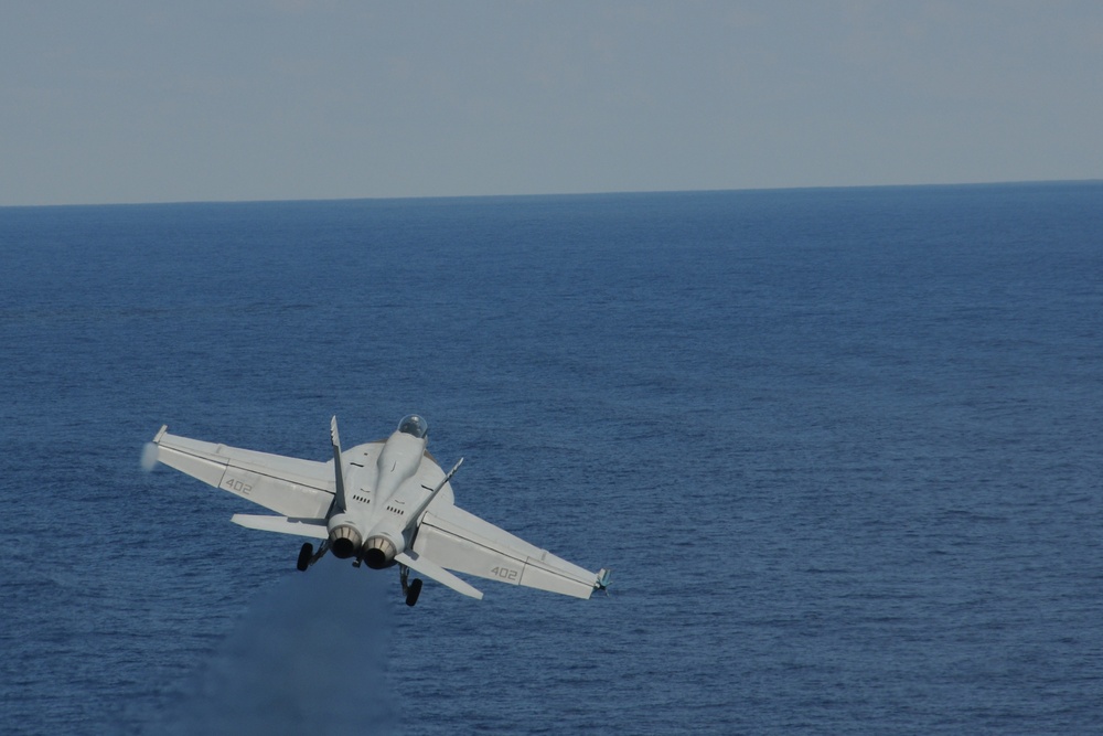 A U.S. Navy F/A-18 Hornet or Super Hornet jet fighter, marked with the number '402', is captured in flight over open ocean. The aircraft appears to be in a banking maneuver, viewed from a slightly elevated rear angle, likely just after launch from an aircraft carrier. The sky is overcast and the sea is calm and deep blue.