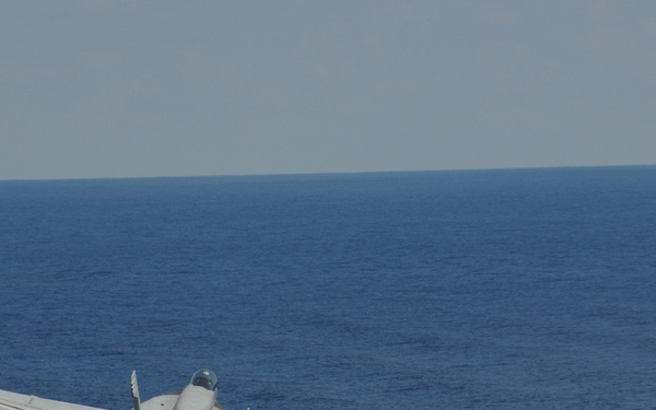 A U.S. Navy F/A-18 Hornet or Super Hornet jet fighter, marked with the number '402', is captured in flight over open ocean. The aircraft appears to be in a banking maneuver, viewed from a slightly elevated rear angle, likely just after launch from an aircraft carrier. The sky is overcast and the sea is calm and deep blue.