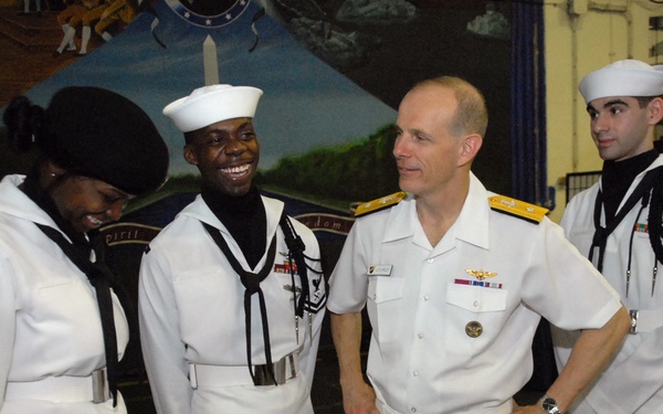 A U.S. Navy officer, appearing to hold a senior rank based on gold shoulder boards and aviator wings, is interacting with enlisted sailors in white dress uniforms. The group appears to be in a hangar bay or large interior space of a naval vessel or installation, decorated with patriotic murals and bunting. The atmosphere is informal and positive, with at least one sailor visibly laughing.