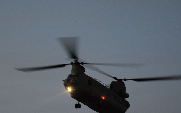 A tandem-rotor military helicopter, consistent with a Boeing CH-47 Chinook, is captured in flight during dusk or dawn conditions. The aircraft is silhouetted against a muted gray-blue sky with its navigation lights and a forward spotlight illuminated. Both rotor systems are visibly spinning, indicated by motion blur on the blades.