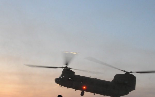 Military personnel in full combat gear observe two tandem-rotor helicopters, identifiable as CH-47 Chinooks, operating at low altitude during dusk or dawn conditions. One helicopter is airborne in the foreground while a second is visible on or near the ground in the background, both kicking up significant dust. The scene appears to depict a military air assault or extraction operation in an arid, desert-like environment.