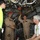 Two military personnel in tan/khaki flight suits are conducting what appears to be a tour or briefing inside a large military aircraft, likely a cargo or patrol aircraft. They are explaining the aircraft's internal systems to two civilians, one wearing a bright yellow shirt and another in a white polo shirt with a lanyard/badge. The overhead area is densely packed with hydraulic lines, wiring bundles, and mechanical systems typical of military aircraft interiors.