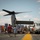 A public open-house or air show event is taking place on the flight deck of a naval vessel, with civilians and military personnel gathered around a V-22 Osprey tiltrotor aircraft. The ship appears to be docked in an urban harbor, with a recognizable city skyline visible in the background. Colorful signal flags are strung along the left side of the deck, adding a festive atmosphere to the military display.