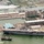An aerial photograph of a naval base showing a large amphibious assault ship docked at a pier, with a historic tall ship sailing nearby flying an American flag. A formation of Blue Angels jet aircraft is performing a flyover, leaving white contrail smoke trails across the sky. The scene appears to be a special naval event or air show, with crowds of people visible on the ship's deck and along the waterfront.