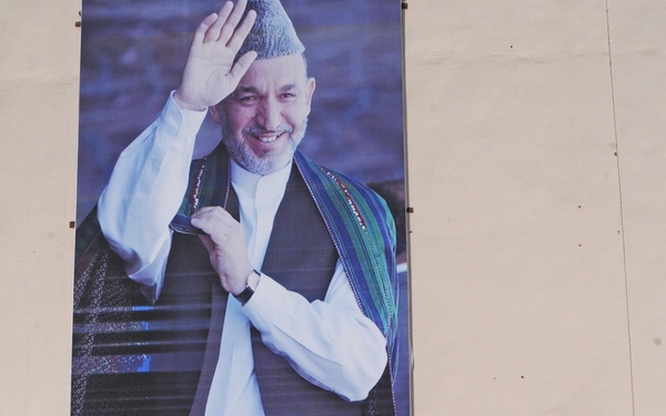 Four individuals stand in front of a large official portrait banner mounted on a building wall, all posing with their arms raised above their heads forming an 'O' shape. The banner contains Dari/Persian script and depicts a smiling man in traditional Afghan attire waving. Three of the four individuals are wearing military uniforms while one is in civilian clothing.
