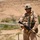 A uniformed military service member stands in an arid, desert environment holding a rifle in a ready position. The soldier is wearing full combat gear including a helmet, tactical vest, and desert camouflage uniform. The background features a sandy hillside, sparse vegetation, and what appears to be a river or water source in the distance.