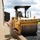 Military personnel in camouflage uniforms are operating a yellow Caterpillar road roller/compactor to pave or compact an asphalt surface adjacent to a building. One service member is seated at the controls of the machine while another stands nearby on a brick walkway, appearing to guide or monitor the operation. The scene takes place at what appears to be a military installation or base facility.
