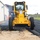 Military personnel are conducting a paving/road construction operation at what appears to be a military installation. A yellow Volvo motor grader (marked 'U.S. AIR FORCE 08D 1072') is spreading fresh asphalt while uniformed airmen assist on the ground. The scene takes place adjacent to a large metal building with additional personnel observing in the background.