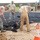 Military personnel in US Air Force ABU (Airman Battle Uniform) camouflage uniforms are performing asphalt/pavement work at what appears to be a military installation. Two workers in the foreground are actively shoveling fresh asphalt, while others observe or supervise. A yellow construction loader/backhoe is visible in the background near a wooden fence perimeter.