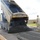 A dump truck is unloading hot asphalt/tarmac material onto a paved surface, with steam/smoke rising from the freshly deposited material. A person in military camouflage uniform and tan boots is observing the operation from the right side. The setting appears to be a military installation, indicated by the uniformed individual and the fenced compound in the background.