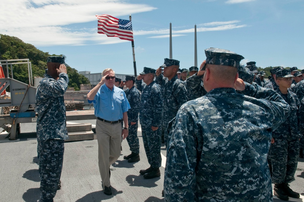 A civilian official in a blue polo shirt and khaki pants walks between two rows of U.S. Navy sailors in digital camouflage uniforms who are rendering a salute. The scene appears to take place on the deck of a naval vessel or pier, with an American flag flying in the background. The setting suggests a formal military inspection, arrival ceremony, or official visit.