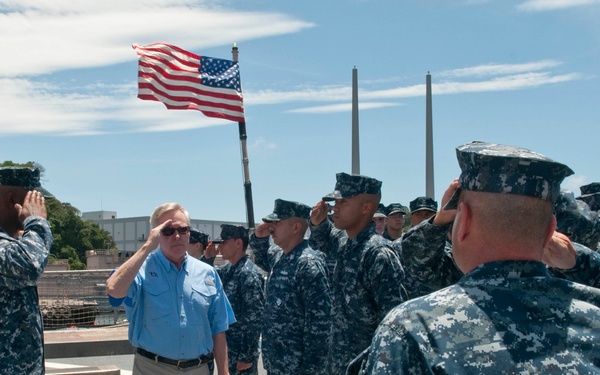 A civilian official in a blue polo shirt and khaki pants walks between two rows of U.S. Navy sailors in digital camouflage uniforms who are rendering a salute. The scene appears to take place on the deck of a naval vessel or pier, with an American flag flying in the background. The setting suggests a formal military inspection, arrival ceremony, or official visit.