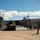 A military amphibious landing craft (LCU or LCAC-type vessel) has beached on a sandy shore, with its bow ramp lowered to offload military vehicles. A service member in olive/green uniform is visible in the foreground, appearing to direct traffic or communicate via radio. The setting appears to be a coastal training or operational area with arid hills visible in the background.