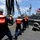 Crew members aboard the USNS Henry J. Kaiser, a U.S. Navy replenishment oiler, are conducting an underway replenishment (UNREP) operation alongside a U.S. Navy warship. Personnel are wearing orange life vests and white hard hats while managing fuel transfer hoses and lines between the two vessels. An American flag is visible flying from the receiving warship in the background.