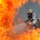 A firefighter in full protective gear, including a breathing apparatus and helmet, is visible through an opening in large, intense flames. The individual appears to be actively engaged in firefighting operations, surrounded by orange and red fire on multiple sides. Smoke and heat distortion partially obscure the figure, creating a dramatic and dangerous scene.