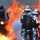 Two firefighters in full aluminized proximity suits and MSA SCBA (Self-Contained Breathing Apparatus) units are facing a large, intense fire. They are holding a red fire hose and appear to be advancing toward or managing the blaze. A fire truck is partially visible in the background to the left.