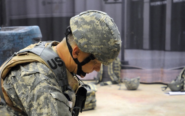 A U.S. Army soldier in ACU (Army Combat Uniform) is performing combat casualty care on another soldier lying on the ground. The standing soldier is leaning over the downed soldier, appearing to assess or treat injuries in what looks like a training exercise. The indoor setting and multiple similar setups visible in the background suggest this is a structured medical training scenario.