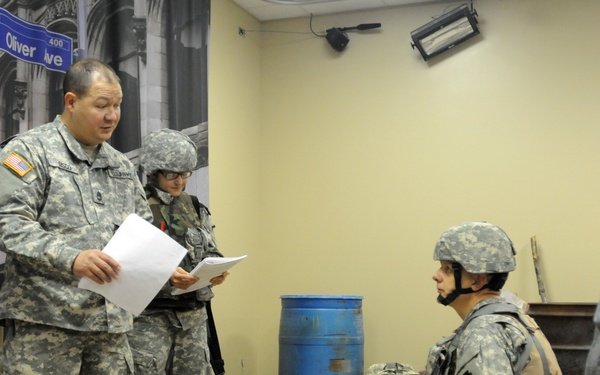 Three U.S. Army soldiers in ACU (Army Combat Uniform) are engaged in what appears to be an indoor training exercise or evaluation scenario. One soldier is standing and reading from papers, likely an evaluator or instructor, while a second soldier kneels beside a third soldier who is lying on the floor, simulating a casualty care scenario. The setting appears to be a mock urban environment used for training purposes.