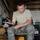 Patriot Exercise 2012 A U.S. Air Force servicemember in ABU (Airman Battle Uniform) camouflage pattern is seated on a rolling stool inside what appears to be a maintenance hangar or workshop. He is working with blue ethernet/network cable, using a crimping tool to terminate a network connector. Various tools and equipment are visible around him on the floor and in the background.