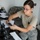 Patriot Exercise 2012 A female military service member wearing US Air Force ABU (Airman Battle Uniform) camouflage pants and a tan t-shirt is seated at a counter or desk. She is holding a mobile phone in one hand and a pen in the other, appearing to transcribe or reference information onto a form. Several rifles or weapons are visible in the foreground on a rack, suggesting this may be an armory or weapons issue point.
