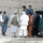A group of approximately 10-12 men are gathered near a mud-brick wall beside a green pickup truck marked 'MP' (Military Police). Several individuals are wearing traditional South Asian/Central Asian attire including shalwar kameez and turbans, while at least one person appears to be in a tactical vest. The setting appears to be a rural or semi-urban environment consistent with Central or South Asian geography.