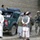 The image shows a military checkpoint or patrol interaction in what appears to be Afghanistan, based on the Afghan flag visible in the background and the mud-brick architecture. A green pickup truck marked '68 MP' (Military Police) is present, with U.S. soldiers in combat gear interacting with local Afghan civilians. The scene depicts a typical civil-military interaction during operations in Afghanistan.