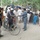 An outdoor scene in what appears to be a rural Afghan village showing a gathering of men, women, and children around a vehicle and bicycle on a dirt road. A uniformed individual, likely a police officer, appears to be distributing or exchanging small items or documents with civilians. The scene suggests some form of community outreach, distribution event, or checkpoint interaction.