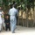 The image shows an outdoor scene in what appears to be a rural Afghan village setting, featuring a group of local civilians interacting with a uniformed Afghan police officer and a fully equipped Western military soldier. A young child is present among the group of adults. The setting includes dirt paths, mud-brick walls, and large shade trees.