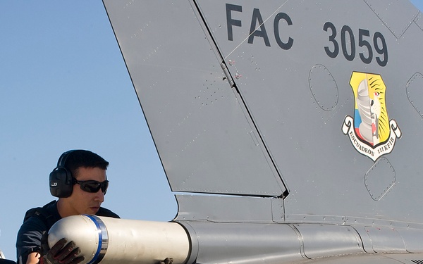 Two military ground crew personnel are working on a military jet aircraft, handling what appears to be a munition or ordnance component near the tail section. The aircraft is marked with 'FAC 3059' and bears a squadron insignia patch, indicating it belongs to the Colombian Air Force (Fuerza Aérea Colombiana). The personnel are wearing hearing protection, sunglasses, and military/ground crew attire.