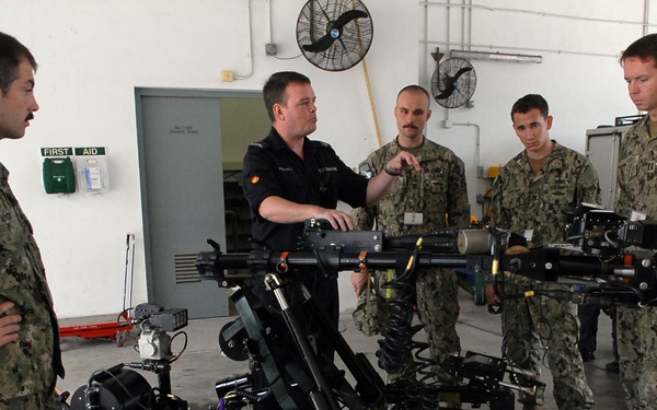 Five military personnel are gathered in what appears to be a maintenance bay or hangar, examining two tracked robotic systems with articulated arms. One individual in a dark uniform, distinct from the others in camouflage, appears to be demonstrating or explaining the robots. The setting suggests a training or familiarization session with EOD (Explosive Ordnance Disposal) or similar ground robots.