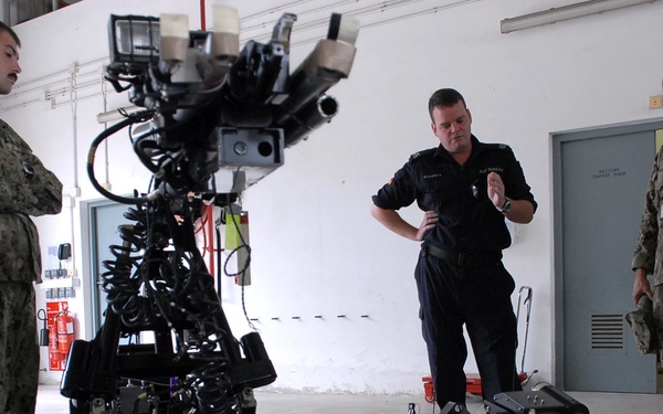 Three personnel are gathered in what appears to be a military or emergency services facility, examining two tracked robotic units on a concrete floor. Two individuals are wearing US Navy digital camouflage uniforms, while a third is dressed in black tactical/emergency services attire. The robots appear to be EOD (Explosive Ordnance Disposal) or remote-operated ground vehicles, with one larger unit featuring a robotic arm and camera system.
