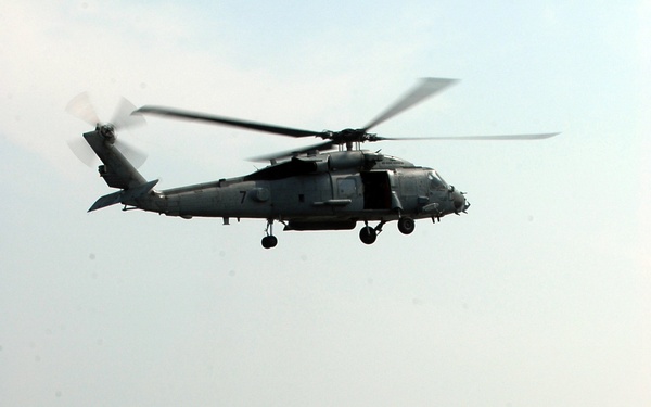 A military helicopter numbered '7' hovers above a Japan Coast Guard patrol vessel designated 'PS 07' operating at sea. The scene appears to depict a joint maritime exercise or coordinated patrol operation between air and sea assets. The image was taken from an elevated vantage point, likely from another aircraft or elevated platform.