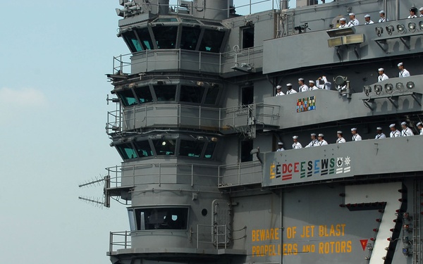 The image shows USS George Washington (CVN-73), a Nimitz-class nuclear-powered aircraft carrier, with hull number 73 prominently displayed on the island superstructure. Sailors in dress white uniforms are manning the rails along the flight deck and on multiple levels of the island, suggesting a formal ceremonial event such as a port arrival or departure. A helicopter, likely an MH-60 Seahawk, is visible in flight to the left of the ship.