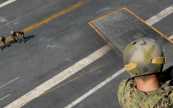 A military personnel member is rappelling or preparing to descend a thick rope from an elevated position, looking down onto what appears to be a large naval vessel's flight deck. Multiple other personnel are visible on the deck below, engaged in various activities. The image is taken from a high vantage point, providing a dramatic top-down perspective.