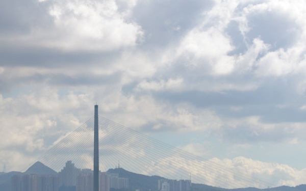 A row of naval personnel dressed in white uniforms stand at attention on a pier or dock, facing a harbor with a prominent cable-stayed bridge and urban skyline in the background. The scene appears to be a formal naval ceremony or 'manning the rails' tradition. The backdrop includes mountains, high-rise buildings, container port cranes, and a partly cloudy sky consistent with a port city in Asia.