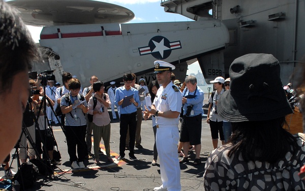 A U.S. Navy officer in dress white uniform is conducting a press briefing on the flight deck of an aircraft carrier. Journalists and reporters surround him, taking notes and recording with cameras and microphones. A military aircraft with U.S. roundel markings and the number 5828 is visible in the background.
