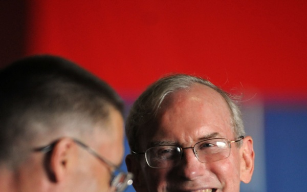 Two men are engaged in conversation at what appears to be a formal or semi-formal event. The man in focus is an older gentleman wearing a dark suit, light blue shirt, and purple patterned tie, smiling broadly. The second man is partially visible in the foreground, out of focus, wearing a white shirt and glasses.