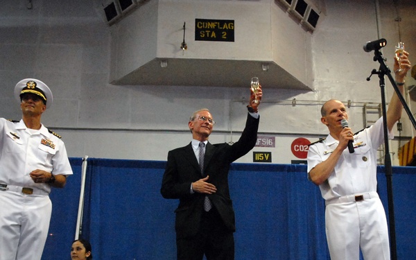 Three individuals stand on a stage raising champagne glasses in a toast during what appears to be a formal naval ceremony. Two men are dressed in white U.S. Navy dress uniforms, while a third man in the center wears a dark business suit. A person in a white chef's uniform stands in the background holding a glass.
