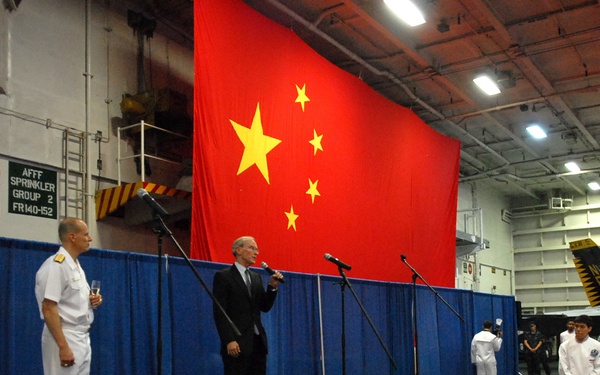 A formal ceremony is taking place inside what appears to be a naval vessel's hangar bay, with a large Chinese national flag (五星红旗) prominently displayed as a backdrop. Two naval officers in white dress uniforms and a civilian in a dark suit stand on a raised stage with microphones, appearing to give speeches or toasts. The setting suggests a diplomatic or bilateral military reception event.