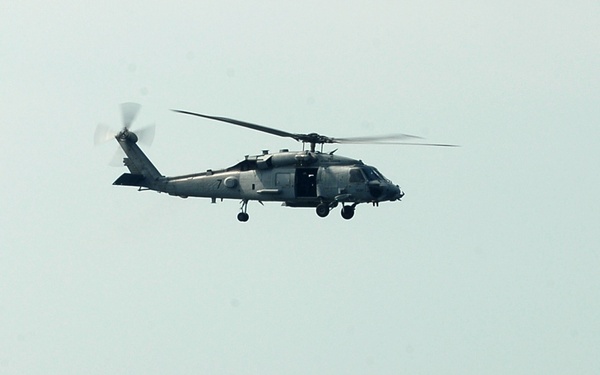 Four U.S. Navy sailors in dress white uniforms stand at attention on what appears to be an aircraft carrier flight deck, observing a military helicopter hovering overhead. The helicopter, consistent with an SH-60 Seahawk variant, is captured mid-flight against a hazy sky. A distant shoreline or cityscape is faintly visible in the background across the water.