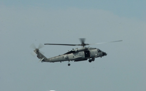 A group of U.S. Navy sailors in dress white uniforms are standing in formation, likely during a ceremony or official event. A military helicopter, appearing to be an SH-60 Seahawk variant, flies overhead in the background. The image captures both the ground personnel and aerial asset in a single frame.