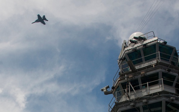 The image is taken from a low angle on what appears to be an aircraft carrier deck, showing a large multi-blade propeller of a turboprop aircraft in the foreground. The carrier's island superstructure with radar domes, antenna arrays, and control tower rises prominently in the background. A jet aircraft, consistent with an F/A-18 Hornet silhouette, is visible flying in the partially cloudy sky.