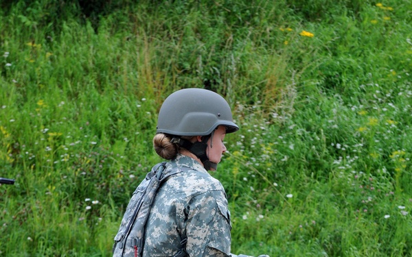 A female soldier in US Army ACU (Army Combat Uniform) digital camouflage stands at what appears to be a firing range, handling a pistol. She is wearing full combat gear including an ACH helmet, body armor/tactical vest, and tan combat boots. A small wooden sawhorse table with ammunition is visible beside her.