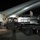 A nighttime ground operations scene at an airfield showing cargo or equipment being loaded onto a large aircraft. A ground support vehicle with a flatbed carrying cargo containers is positioned adjacent to the aircraft's open cargo door. One person is visible standing on top of the cargo load, apparently supervising or directing the loading operation.