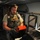 A military or tactical personnel member identified by a name tape reading 'THOMPSON' is standing inside what appears to be a military aircraft, likely a medevac or cargo aircraft. He is wearing a plate carrier vest, tactical gloves, and aviation headset while operating an orange-cased tablet device. The interior shows medical/cargo equipment, litter brackets on the wall, and an illuminated cockpit visible in the background.
