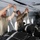 Two individuals are working inside what appears to be a military cargo aircraft, securing loads to an overhead anchor line or static line cable. One person is in civilian-style military attire (tan t-shirt) and the other is in full military camouflage uniform. They are attaching straps or deployment lines to the overhead cable above a stack of black cargo bundles.