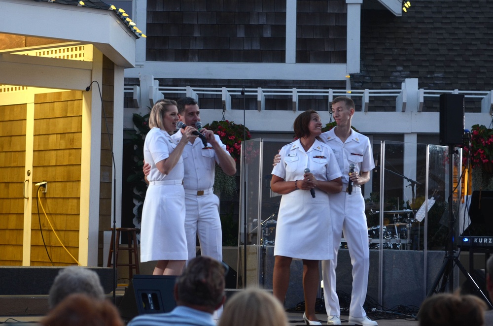 Four individuals dressed in white military-style uniforms are performing on an outdoor stage, with two of them holding microphones and appearing to sing. The performance is taking place at what appears to be an outdoor venue with a shingled building in the background and a band setup visible behind them. An audience is visible in the foreground watching the performance.